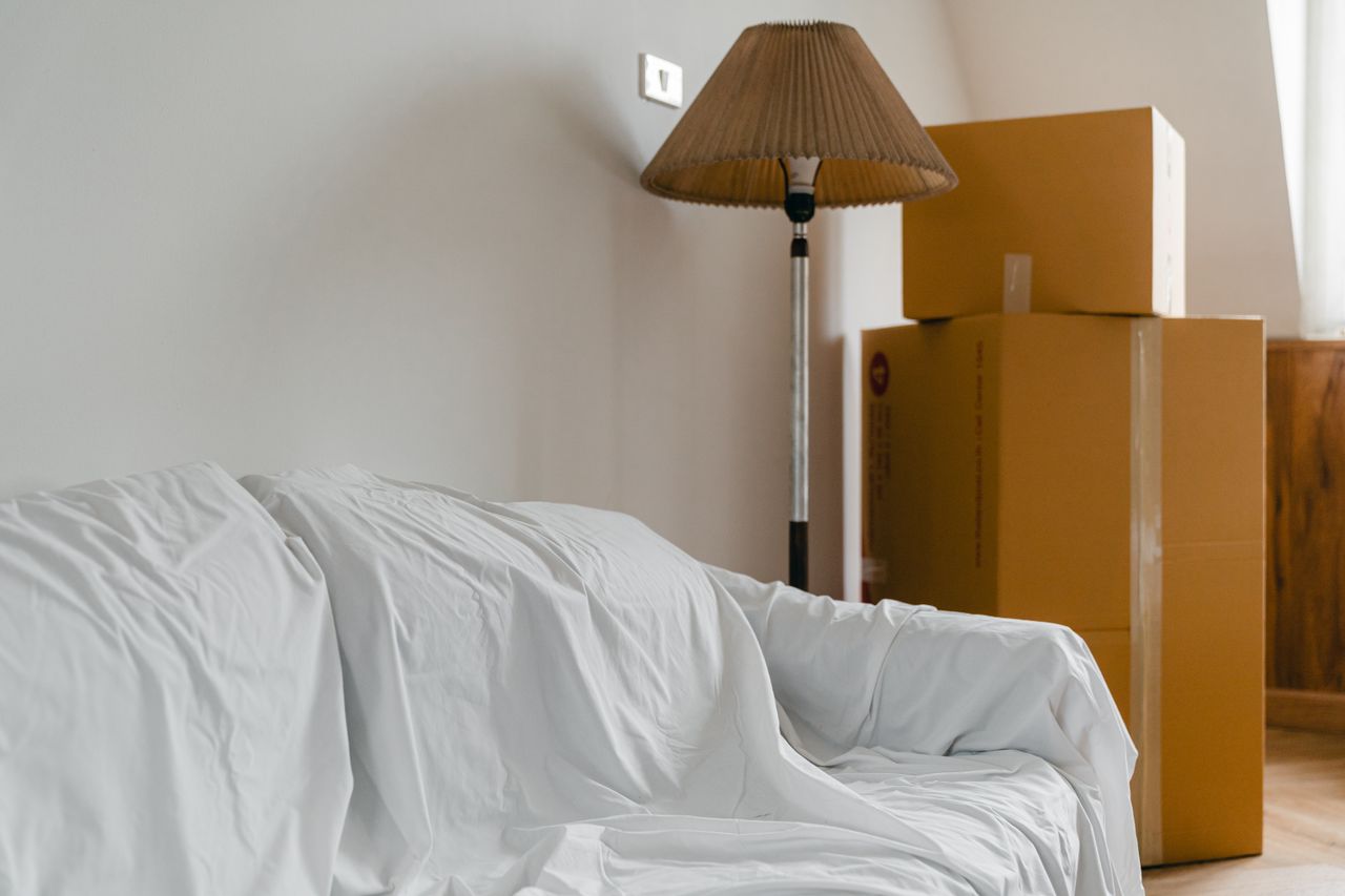 hero-img-02 Covered with sheet sofa and floor lamp and packed cardboard boxes in light spacious room of new contemporary house